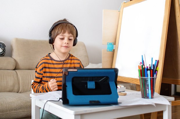 Happy little boy attending online classes from home. Child using digital tablet and headsets for connecting with teacher and schoolmates. School education and social distance during quarantine