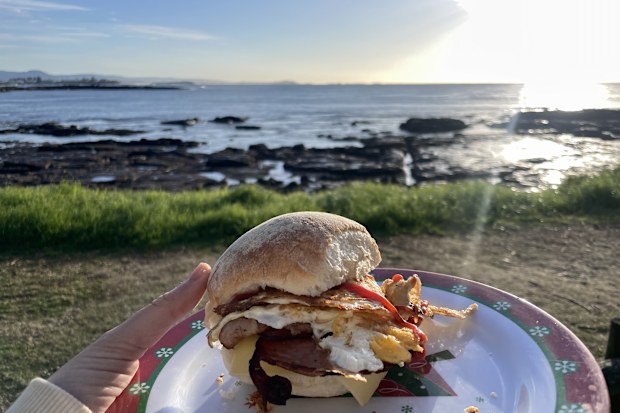 A perfect bacon and egg roll on a plate, with a beach in the background