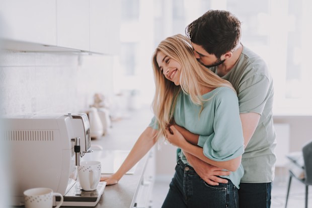 Bearded guy standing behind blonde girl and kissing her. They are waiting for hot drink