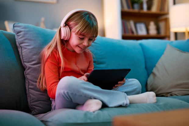 Smiling little girl is using a digital tablet and wearing headphones while sitting comfortably on a sofa in her living room, enjoying her leisure time at home