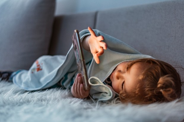 Close up of adorable kid laying down on the bed by holding smartphone at night. Photo of Little kid in bed under a blanket looking at the smartphone at night. Happy smiling baby boy laying on his tummy on the bed under a blanket and plays on a smartphone in a game in the dark. The child's face is illuminated by a bright monitor.