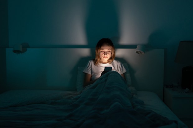 Sleepy exhausted woman lying in bed using smartphone, can not sleep. Insomnia, addiction concept. Sad girl bored in bed scrolling through social networks on mobile phone late at night in dark bedroom.