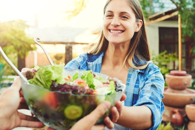 Shot of young woman taking a salad bowl from a friend while sitting around a table outdoors