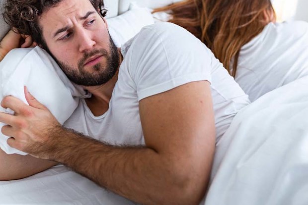 Man and woman lying in bed. The man is in front and looking suspicious of the wife, who is turned the other way, asleep.