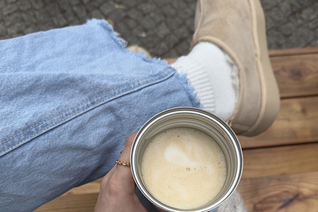 Personal perspective of woman holding a latte while sitting on a wooden bench in light blue jeans and cozy sweater