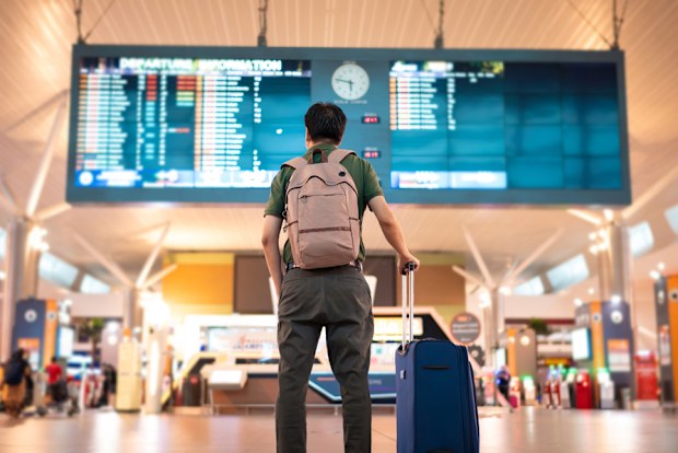 Asian male traveler with wheeled luggage checking for flight schedule on arrival and departure board at Kuala Lumpur International Airport