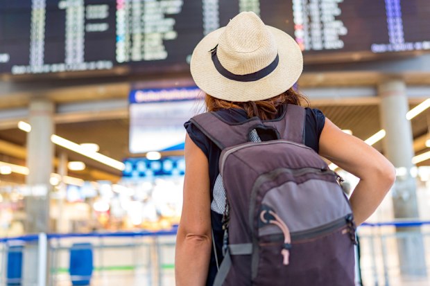 A woman wearing a straw hat and a backpack is standing in front of a large airport display. She is waiting for her flight, possibly at an airport