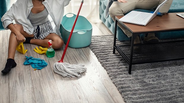 Woman cleans the floor at home while man sits on the couch and uses laptop