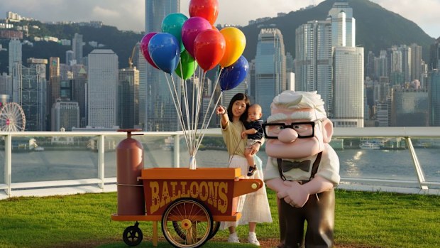 A woman holding a baby poses for photos next to a sculpture of Carl Fredricksen from 'Up.'