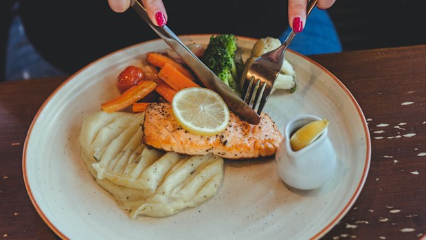 A woman sitting at the table in a cafe, eating redfish steak and mashed potatoes with vegetables on a white plate