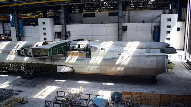 The massive unfinished fuselage towers over everything in the giant warehouse, with its wings and other parts stored nearby. 