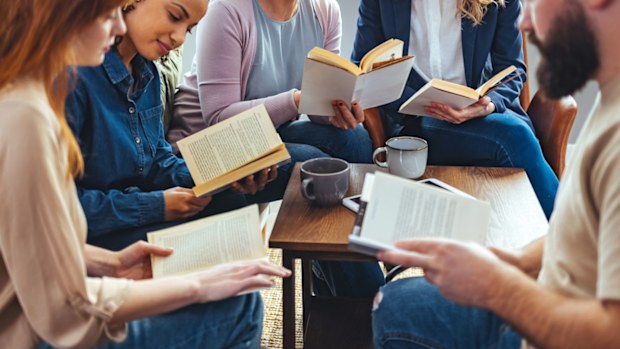 STOCK IMAGE: Small group of people with a mixed age range sitting at a table, discussing and reading books together.