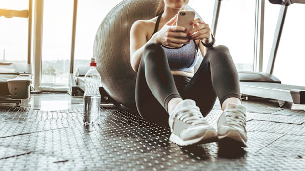 Young woman athlete using cell phone at gym. Young woman in sportswear checking phone while resting after workout on floor. Beautiful fit girl messaging with smartphone at fitness centre.