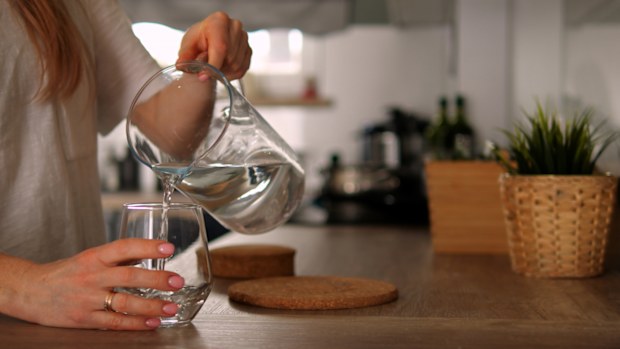 Faceless young woman pour pure water from jug to glass in kitchen. Anonymous girl in white t-shirt holds jar takes care of water balance