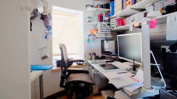 A shot of a messy desk in a home office, the room is small and cluttered.