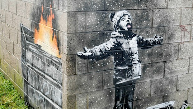 An artwork by Banksy on the side of a garage depicts a child dressed for snow playing in the falling ash and smoke from a skip fire, in Port Talbot, Wales.