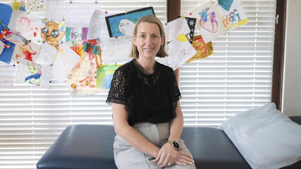 Dr Rebekah Hoffman sits on a medical bed at her Sydney practice. She's wearing grey trousers, a black shirt, and there are children's drawings on the wall behind her.