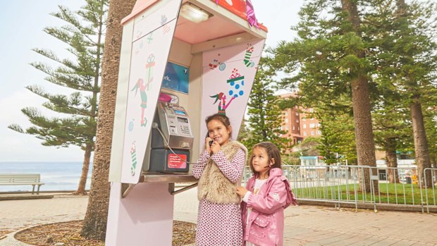 Two little girls standing at a pay phone, the older one is talking into the receiver.