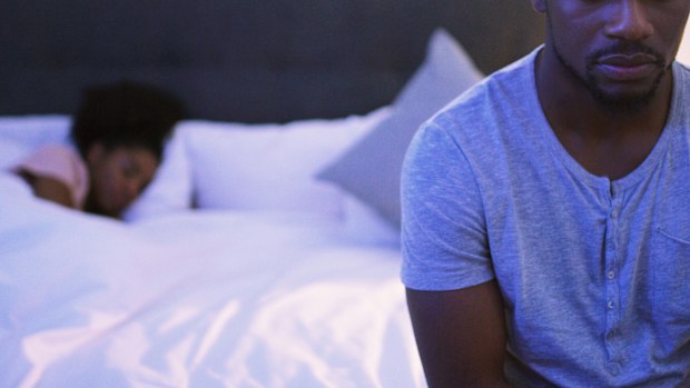 Shot of a concerned looking young man sitting on a bed while his wife sleeps in the background
