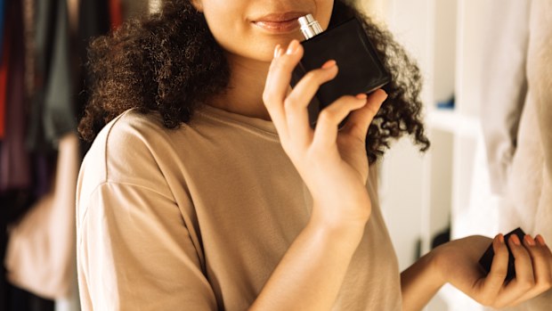 Close-up of charming afro american teenager holding glass bottle and sniffing perfume. Attractive African girl in casual clothes has closed her eyes and is enjoying the scent of perfume. Modern interior.