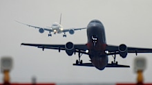 A Qantas Boeing B787-9 plane, registration VH-ZNL, coming into land from the north on the main runway of Sydney Kingsford-Smith Airport as flight QF28 from Santiago.  In the foreground is a Jetstar A320-232 plane taking off from the main runway and heading to the Gold Coast as flight JQ422.  In the near foreground are landing lights and their structures. This image was taken near Nigel Love Bridge, Mascot on an overcast and rainy afternoon on 25 November 2023.