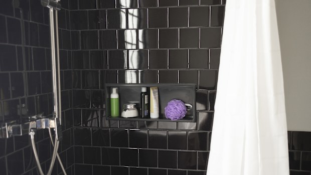 A shot of a black-tiled bathroom with bathroom toiletries on a marble shelf and a shower faucet and a curtain.
