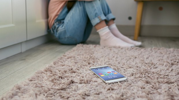 Mobile phone lying on the floor with unrecognizable sad little girl sitting behind. Selective focus on mobile in foreground
