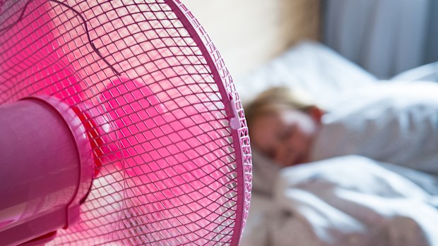 A young girl sleeps in her bed and has a pink fan on her desk blowing cool air over her.