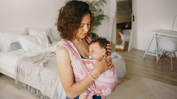 Casually dressed young mixed race woman with wavy hair spending day at home, cradling her infant child, wearing baby carrier. Hispanic mother watching her adorable son sleeping in wrap sling