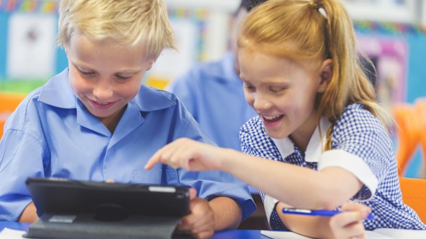 Students using a digital tablet in a classroom. There is a boy and a girl, both are wearing school uniforms and are happy and smiling.