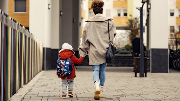 A young woman, mother of nanny, holding hands with little three year old boy with a schoolbag and taking him to kindergarten.