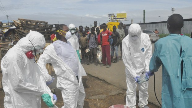 Onlookers watch as health workers in protective gear spray each other with disinfectant in Monrovia, Liberia. (AAP)