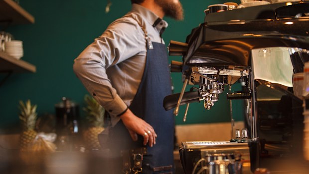 Side view of unrecognizable barista standing behind bar counter, hands on hips, taking customer's order, espresso machine ready to go.
