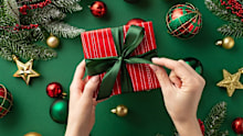 Christmas concept. First person top view photo of woman's hands untying ribbon bow on red giftbox over gold green baubles star ornaments mistletoe pine branches in snow on isolated green background
