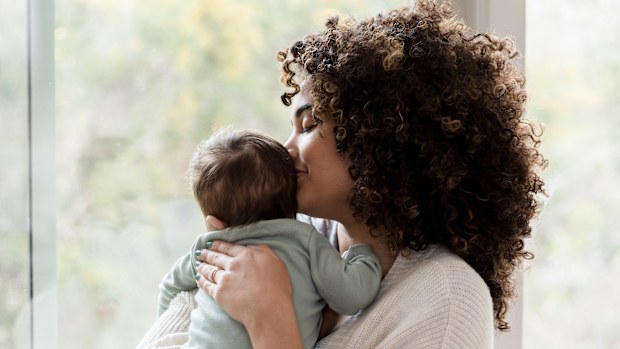 Young mom gives her baby a kiss on his head while standing next to a window in their home
