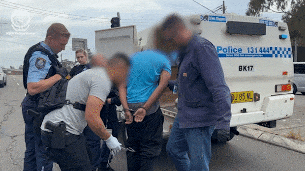 A man in a blue shirt with his face blurred being escorted into a police car in handcuffs.