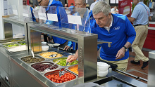 President Thomas Bach tries food from a salad bar while touring the Olympic Village.