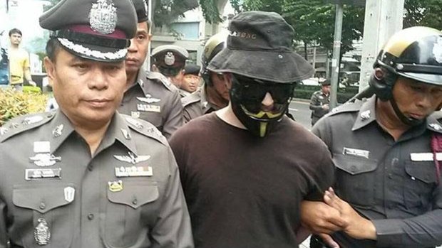 A man in a Guy Fawkes mask and sunglasses is escorted by Thai police in Bangkok. 