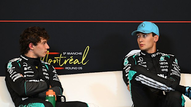 Two men sit on a couch in F1 suits. 