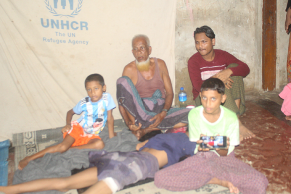 Mohammed Kasim (centre, in singlet) with family members, including 12-year-old Mohammed Hubaib (sitting at the front),  by a UNHCR banner at their shelter in Teknaf, Bangladesh. The boy lying down is so ill he cannot move and has urinated and defecated in his clothes. 