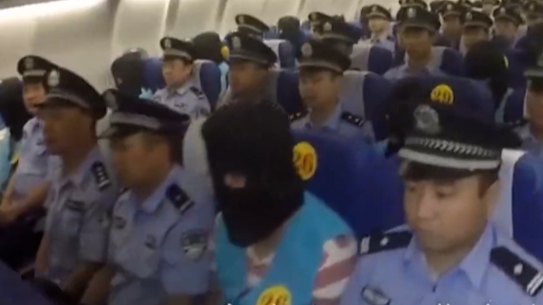 A hooded person, suspected of cyber scams, sits  between Chinese police officers on a charter plane from Fiji to China in 2017.