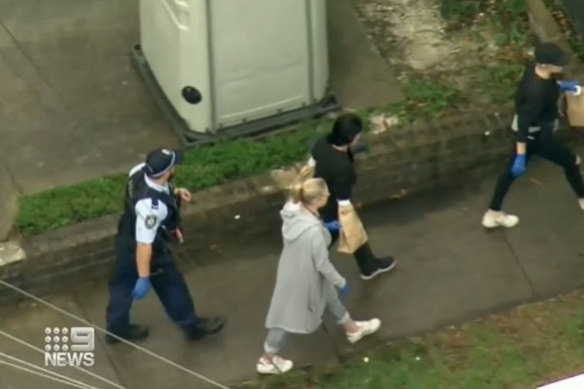 The woman is escorted by police, with her hands in evidence bags.