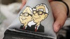 A clerk displays a souvenir phone holder with illustrations of CEO Warren Buffett (right) and vice-chairman Charlie Munger. Asked to explain Berkshire Hathaway’s failure to best its own yardstick, Munger said “If this is failure I want more of it.”
