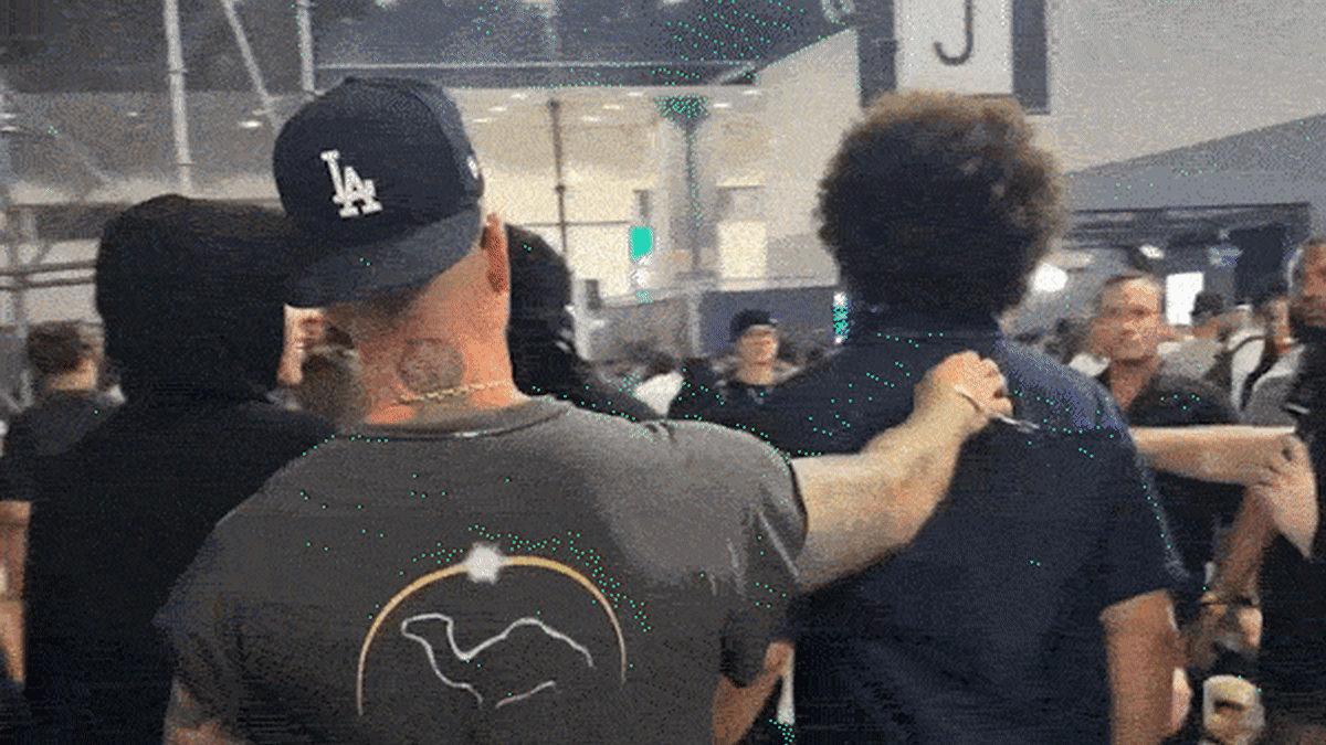 People in dark clothing at indoor venue confrontation scene, one wearing LA Dodgers cap