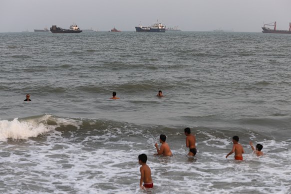 Iranian children swim in the Strait of Hormuz as cargo ships are anchored in the distance.