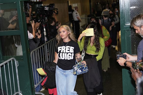 Members of the public, including a woman wearing a "Free Luigi" shirt, enter a courtroom before Luigi Mangione is escorted in.