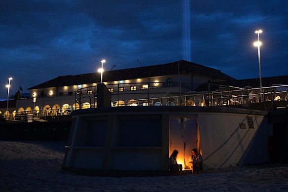 People light candles while light beams are projected from Bondi Pavilion to mark a week since 15 people were killed in the Bondi Beach massacre.