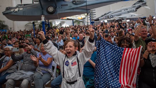 A young boy wears an astronaut costume next to a woman waving a flag as they watch a live broadcast of the return of the Artemis II crew.