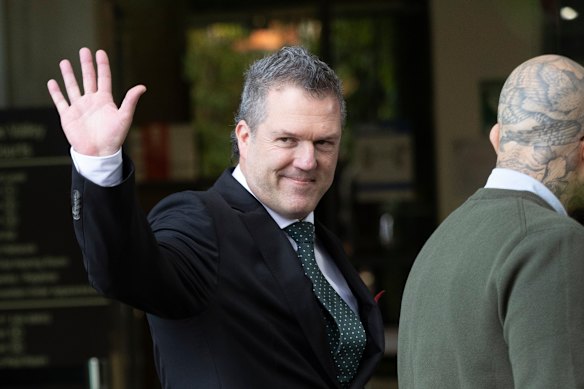 Dr Chris Webster waves to the cameras in his Hugo Boss suit outside court.