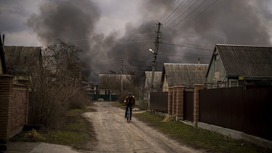 A Ukrainian man rides his bicycle near to a factory and a store burning after been bombarded in Irpin, on the outskirts of Kyiv, Ukraine, Sunday, March 6, 2022. (AP Photo/Emilio Morenatti)